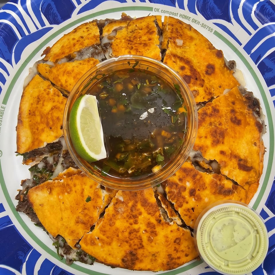 A large, golden-brown quesadilla, cut into wedges, with a side of dark dipping sauce and a lime wedge.