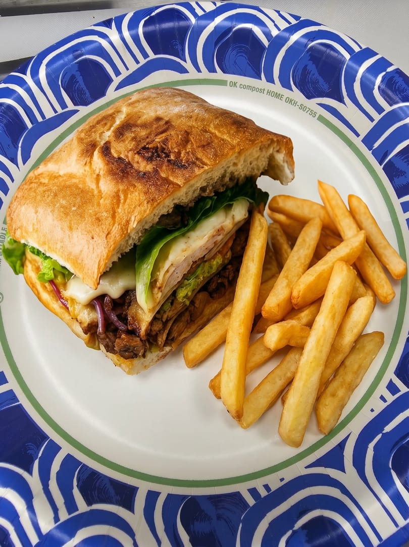 A chicken sandwich with melted cheese, lettuce, and onions, served with golden french fries on a paper plate.