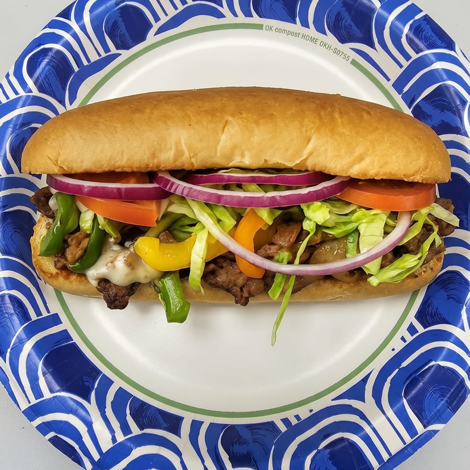 A steak and cheese sub with lettuce, tomato, onions, and peppers on a paper plate.