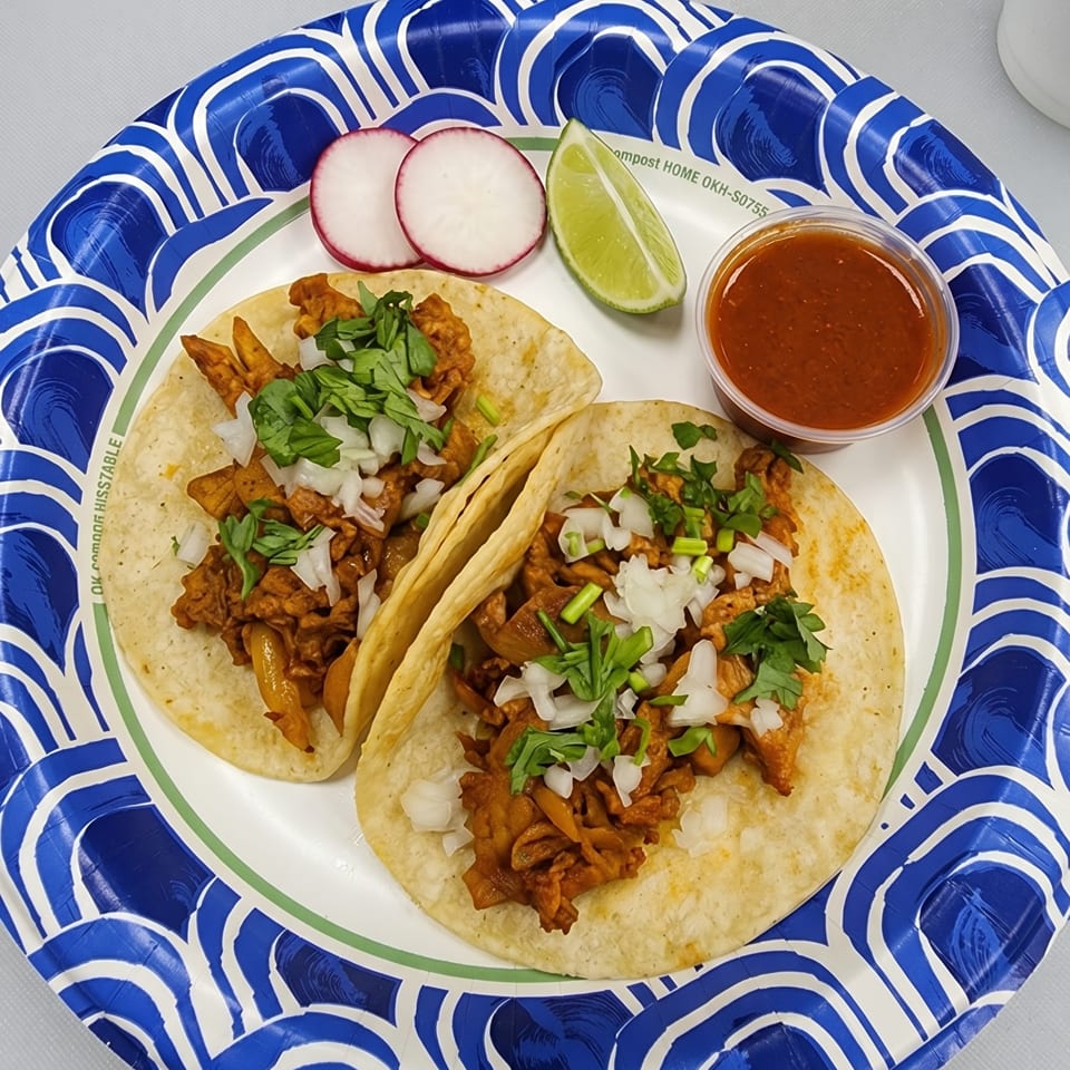Two tacos with meat, onions, and cilantro, served with radishes, lime, and salsa on a paper plate.